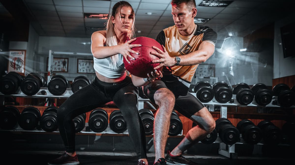 A man and woman perform a tandem workout with a medicine ball in a gym setting.